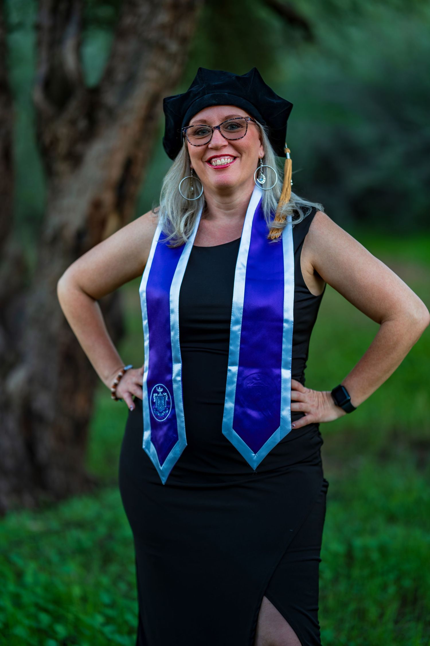 Graduate wearing black dress and blue graduation stoles poses confidently outdoors.