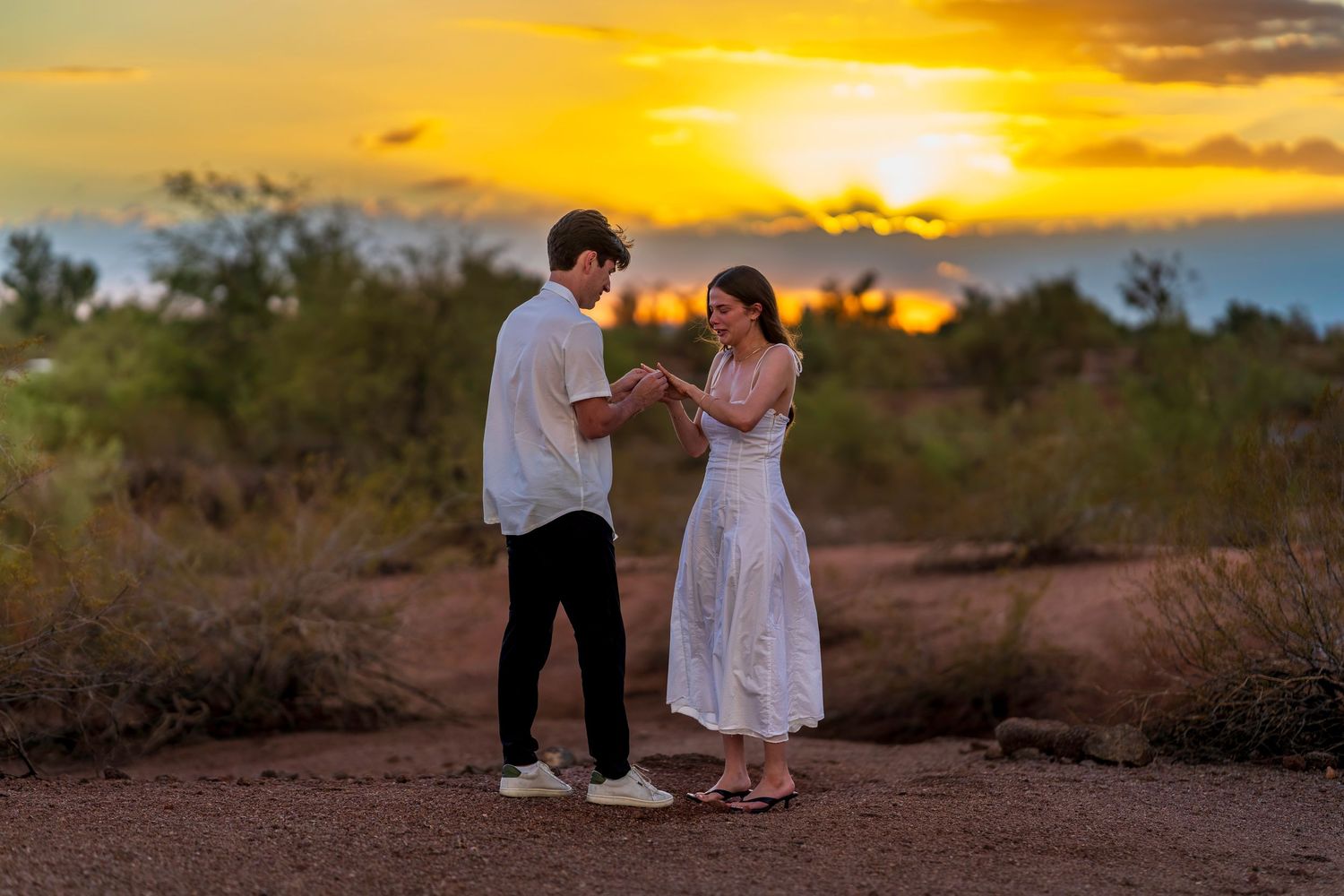 A romantic desert photoshoot with a couple silhouetted against a vibrant orange sunset sky.