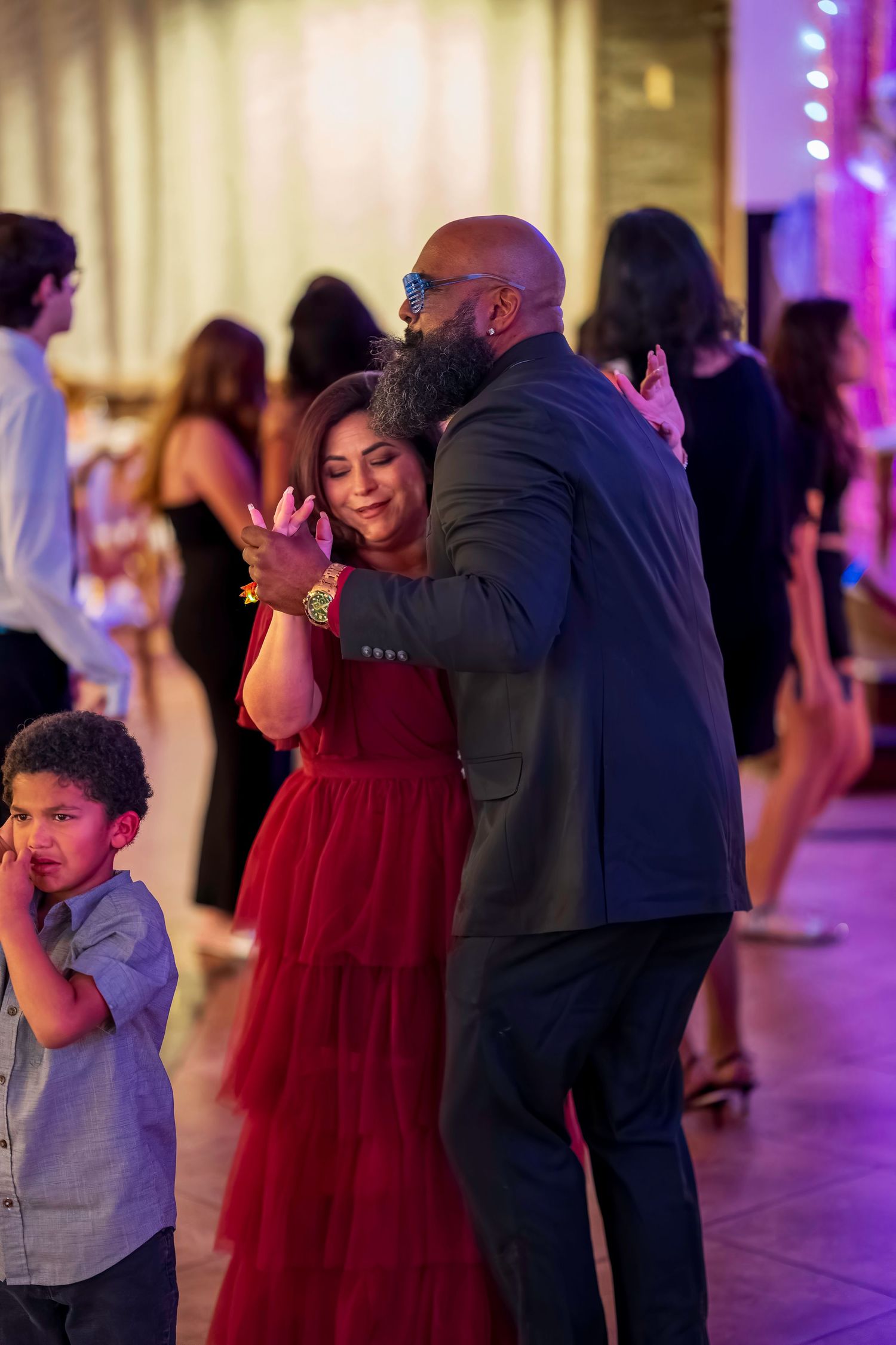Couple dancing closely at a festive event, woman in red dress, bearded man in suit, child nearby, colorful lights.