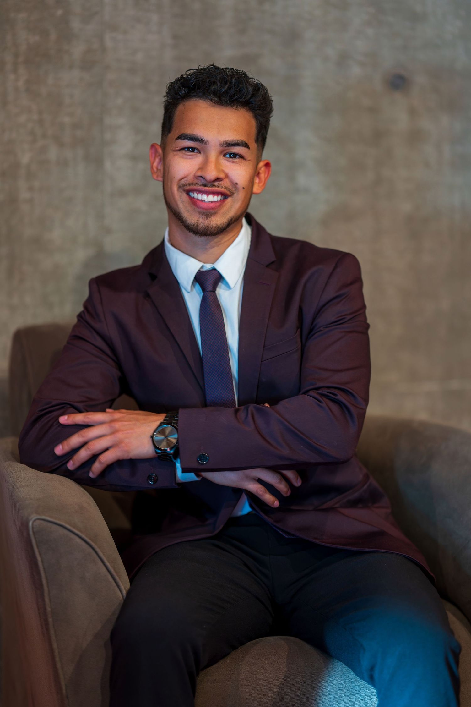 Professional portrait of person in burgundy suit seated on leather chair.