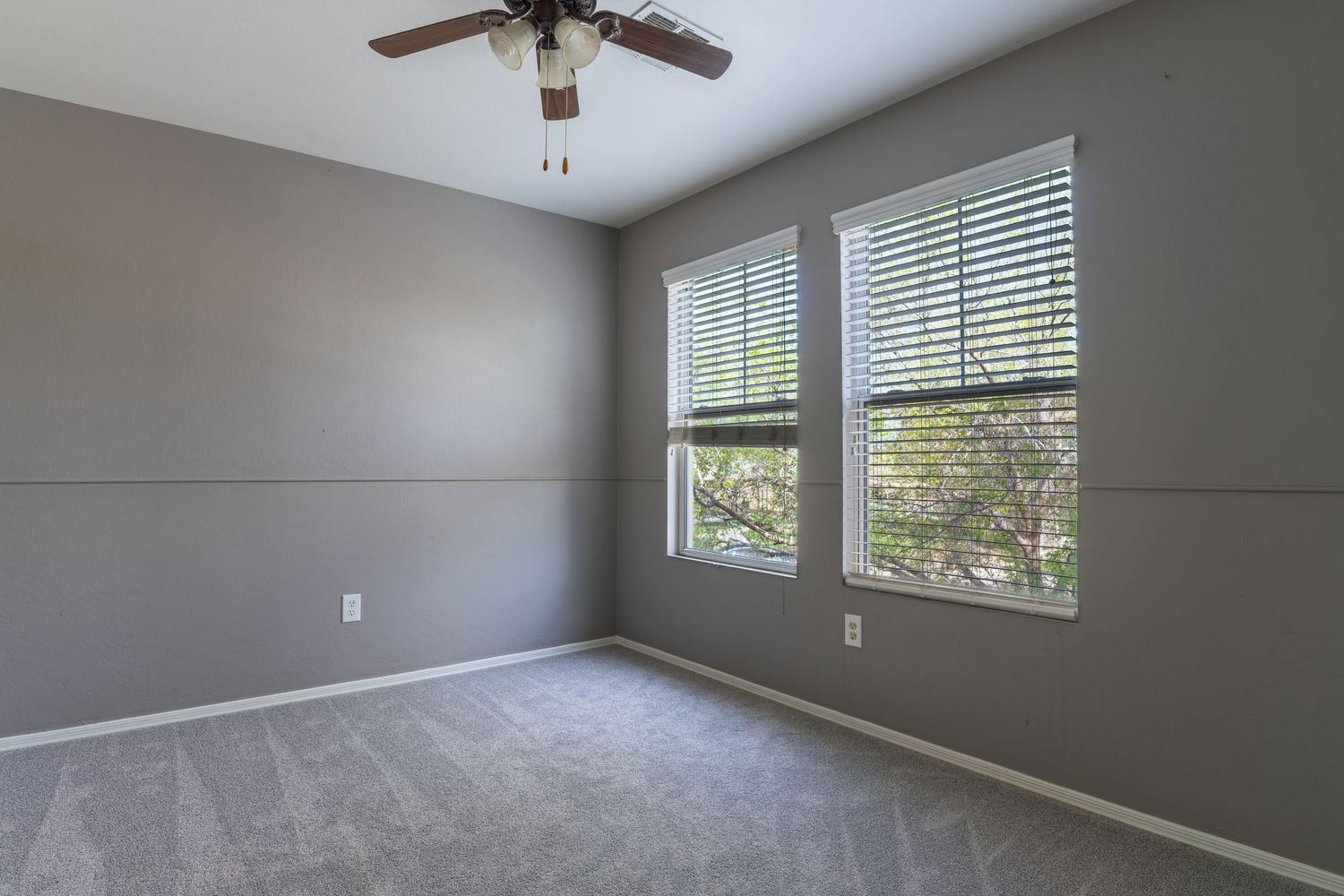 Gray carpeted bedroom with ceiling fan, two windows with white blinds, and neutral gray walls.