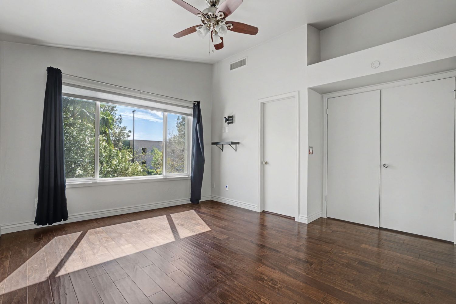 Spacious bedroom with dark hardwood floors, ceiling fan, large window with curtains, and white closet doors.