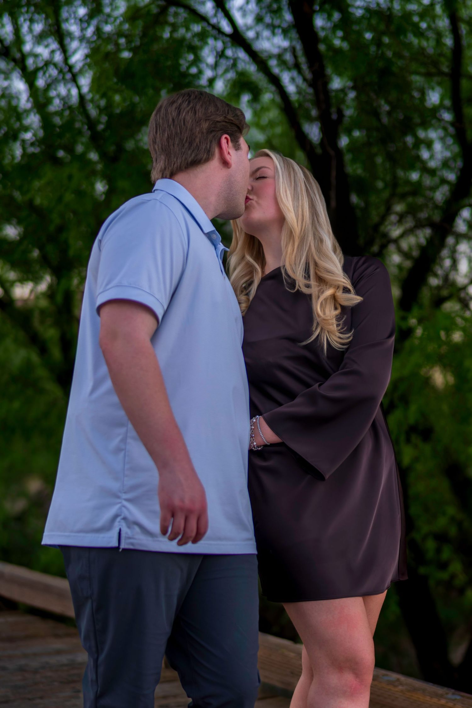 Couple sharing a tender kiss on a wooden bridge surrounded by lush green trees at dusk.