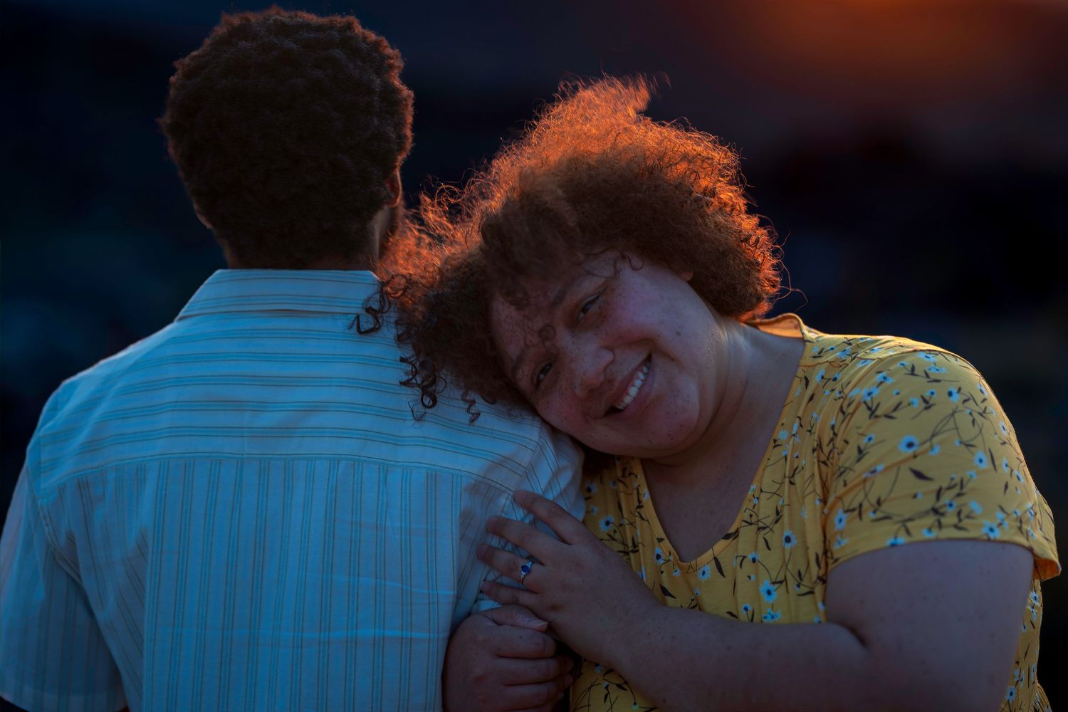 A woman with curly hair in a yellow dress smiles while embracing a man at sunset.