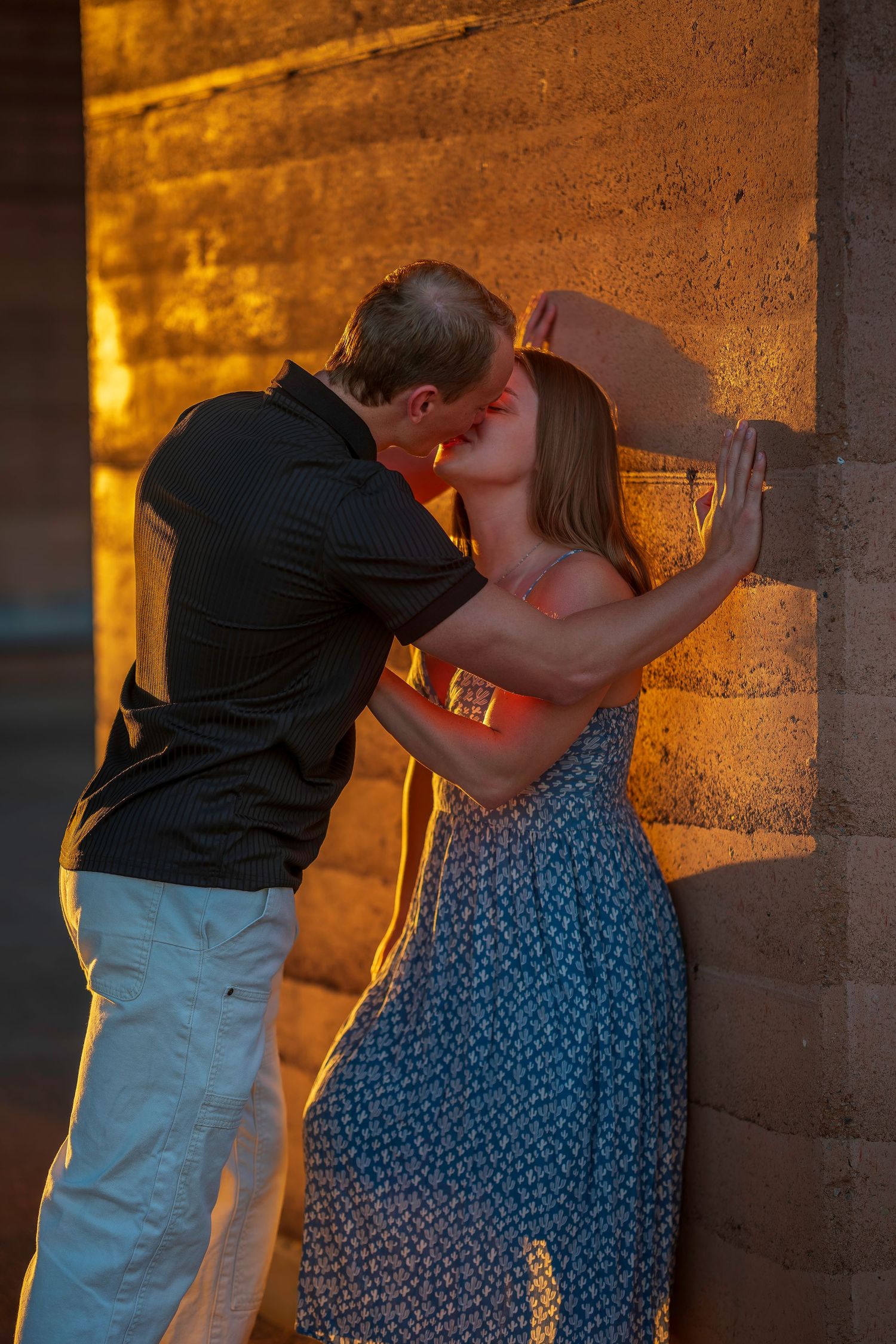 A couple shares a romantic kiss against a warmly lit stone wall at sunset, she wears a blue floral dress.