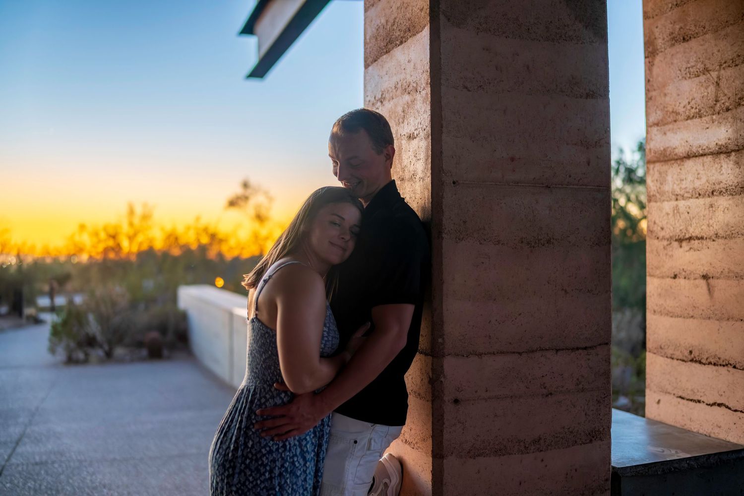 A couple shares an intimate embrace by a stone pillar at sunset.