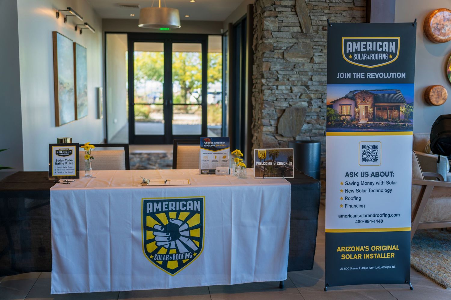 Display booth for American Solar Roofing company with white tablecloth and stone wall backdrop.