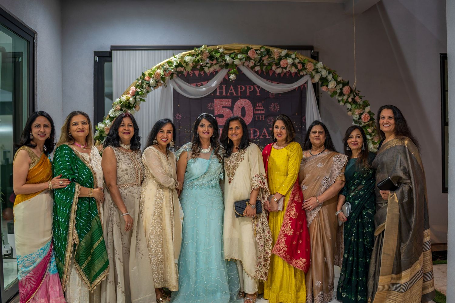 A group of women in colorful traditional attire pose under a floral arch at a celebration.