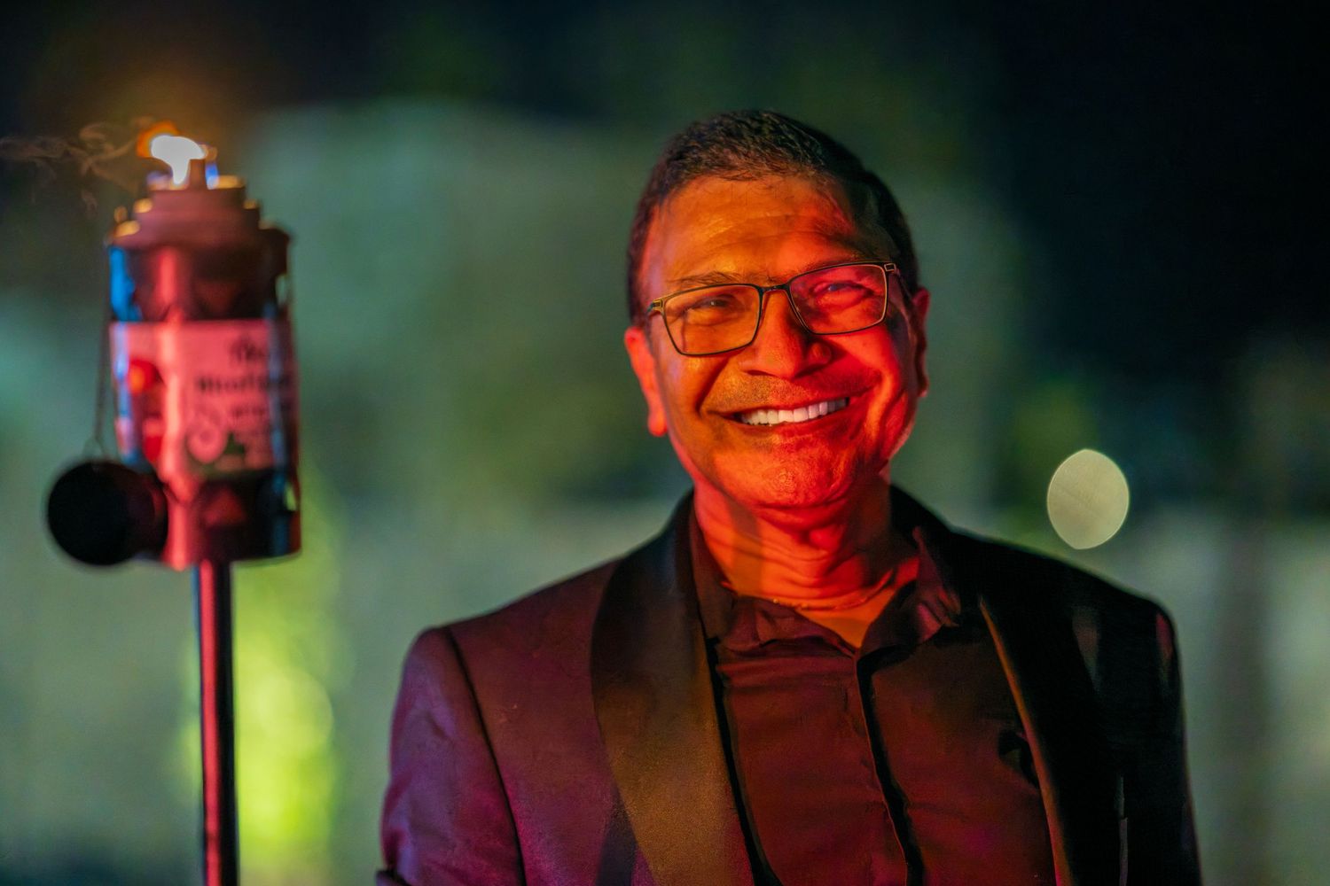 A person in a dark suit smiles warmly while holding a sparkler in dramatic lighting.