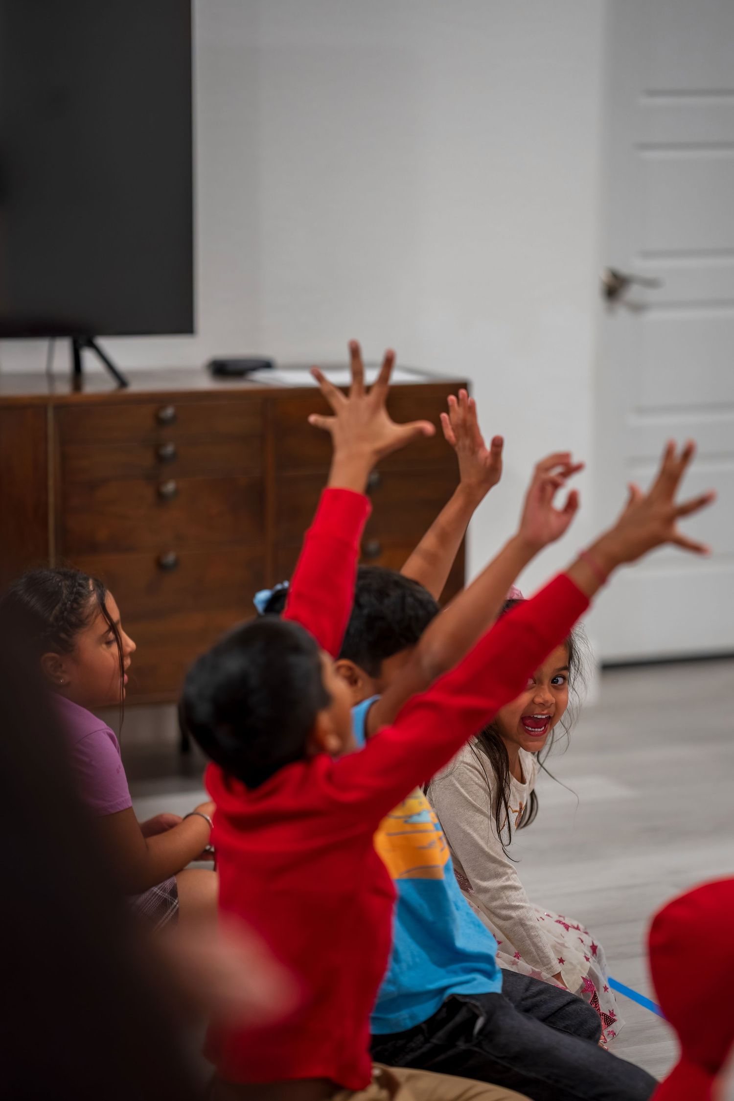 Children raise their arms during an energetic dance or movement activity against a white wall background.
