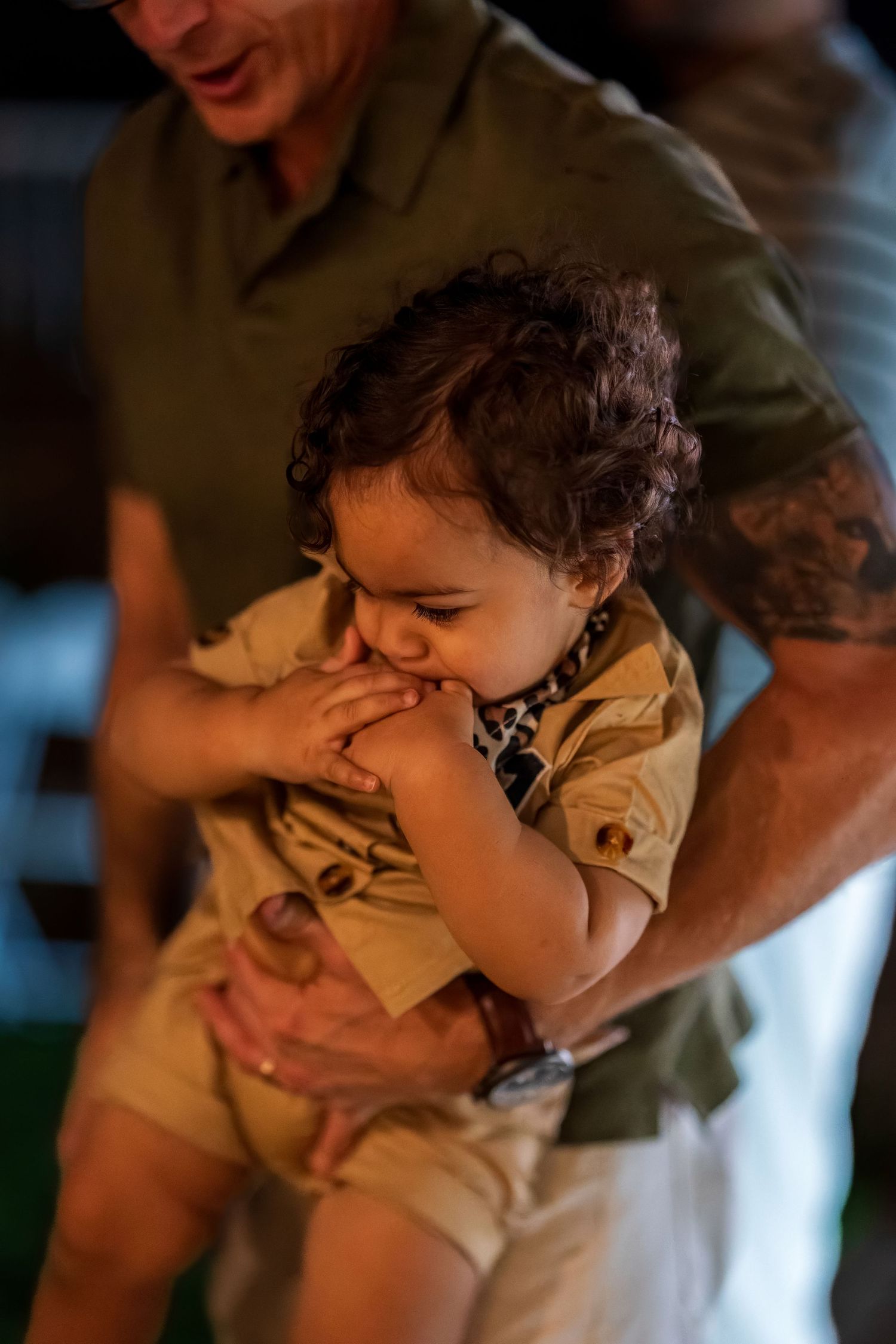 A toddler in a tan outfit chewing on their fingers while being held.