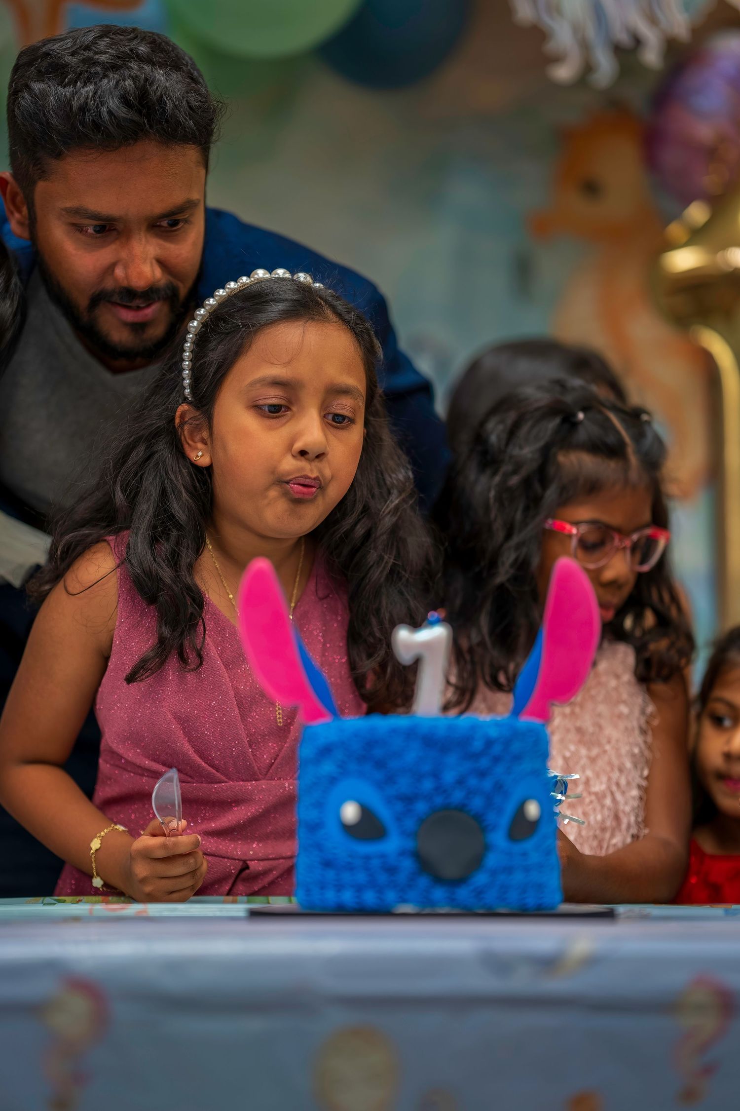 A young girl blows out a candle on a blue Stitch-themed birthday cake at a party.
