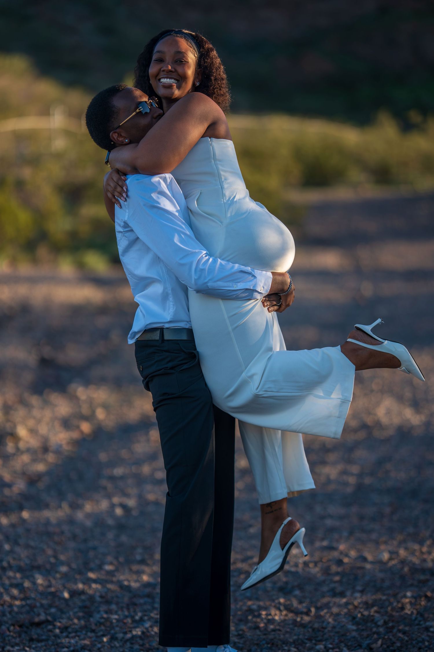 A joyful groom lifts his smiling bride in a white dress outdoors during golden hour.