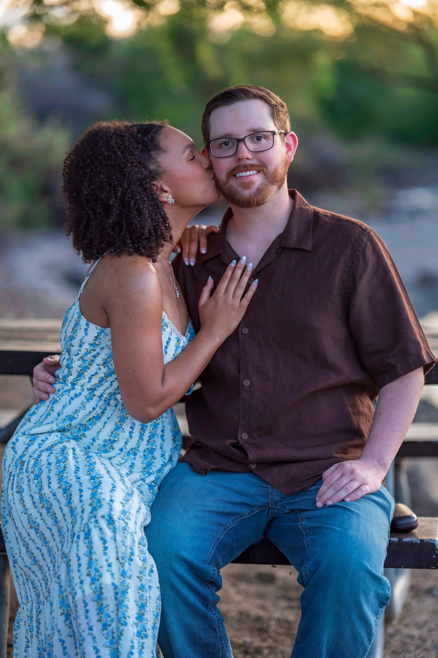 A woman in a floral dress kisses a smiling bearded man with glasses on the cheek outdoors.