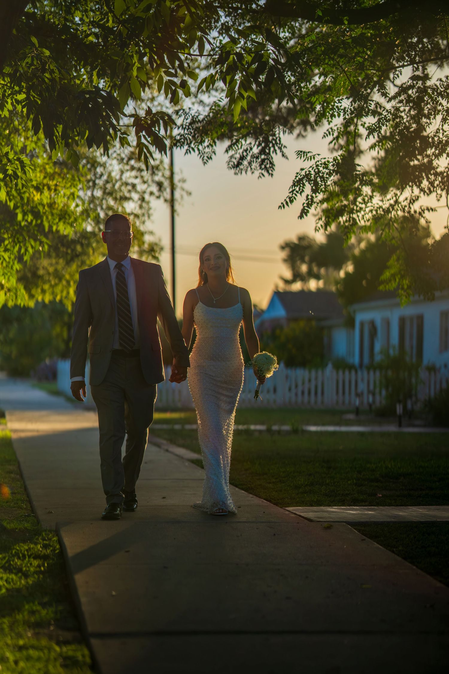 Bride and groom walking hand in hand on a sidewalk at golden hour sunset, framed by lush green trees.