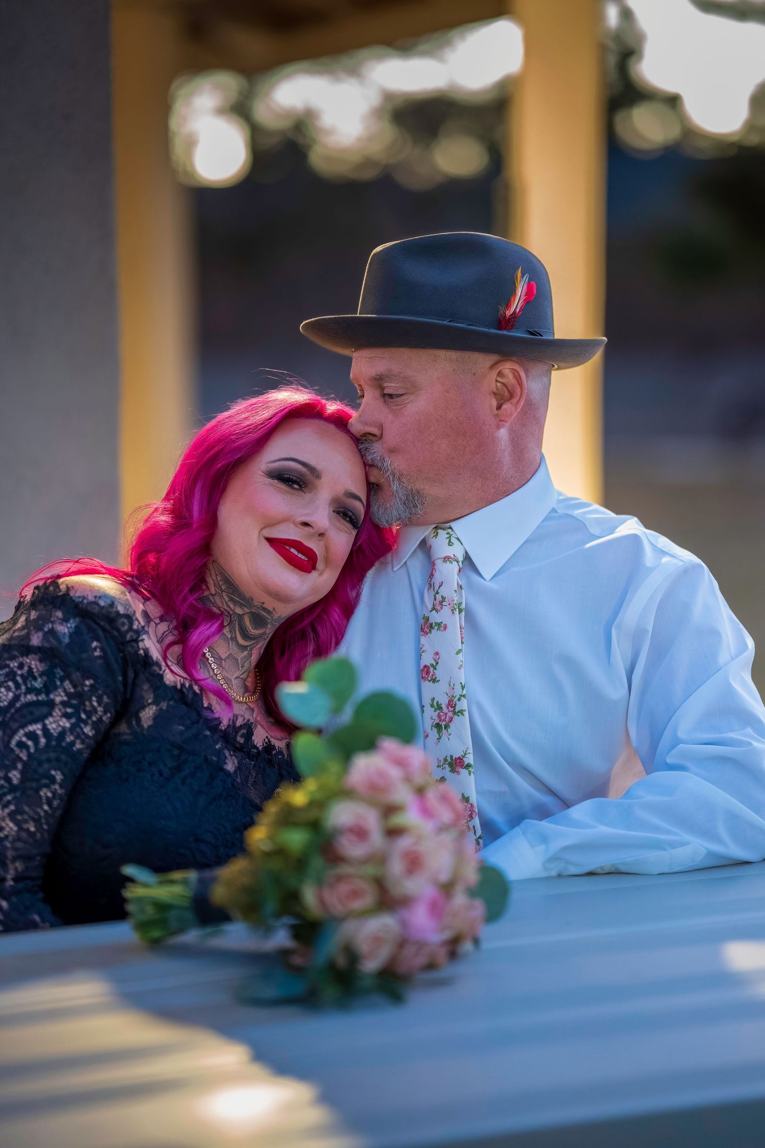 A couple shares a romantic kiss at a wedding, woman with pink hair and tattoos, man in gray hat and floral tie.