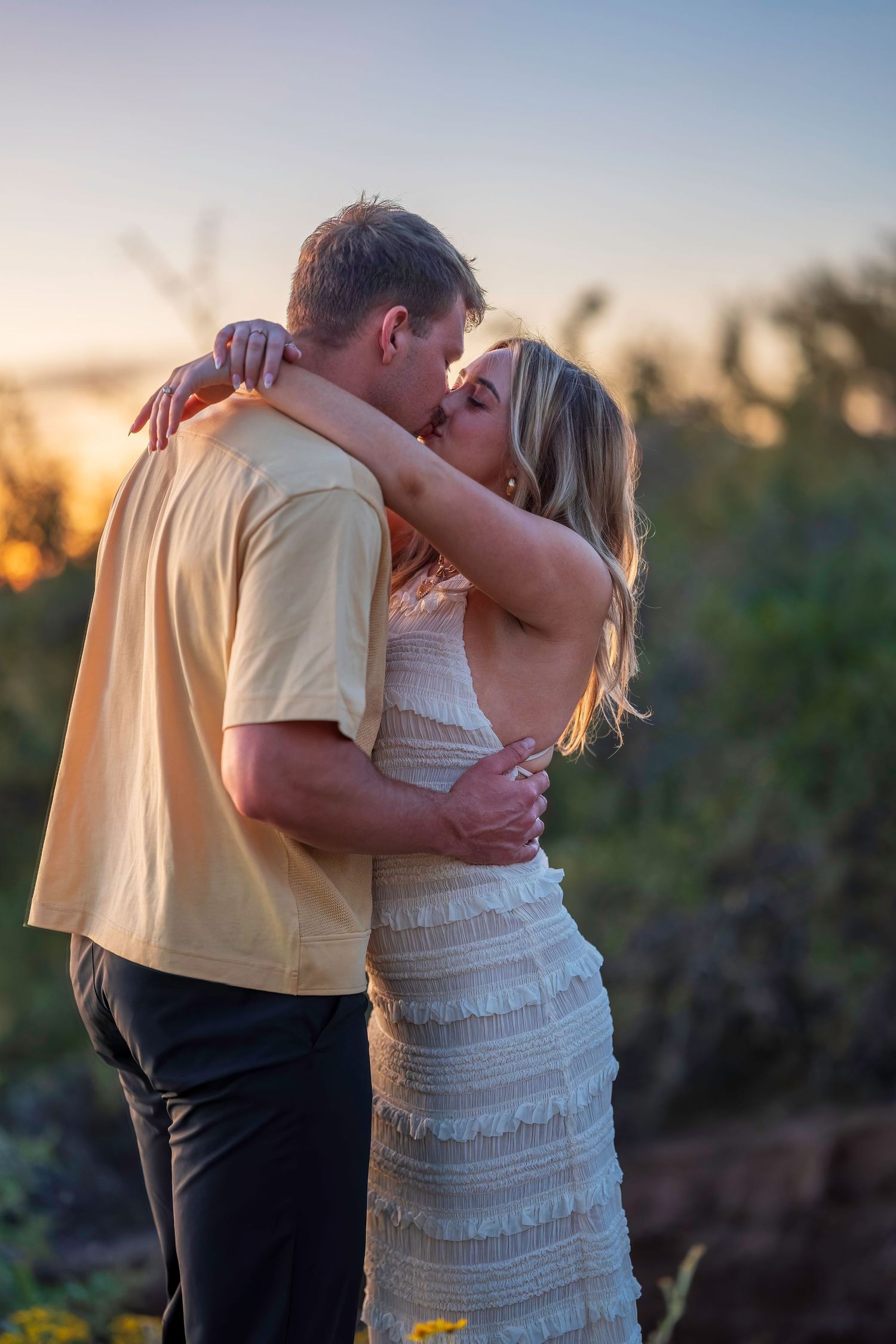 Couple sharing a romantic kiss outdoors during golden hour sunset in a natural landscape.
