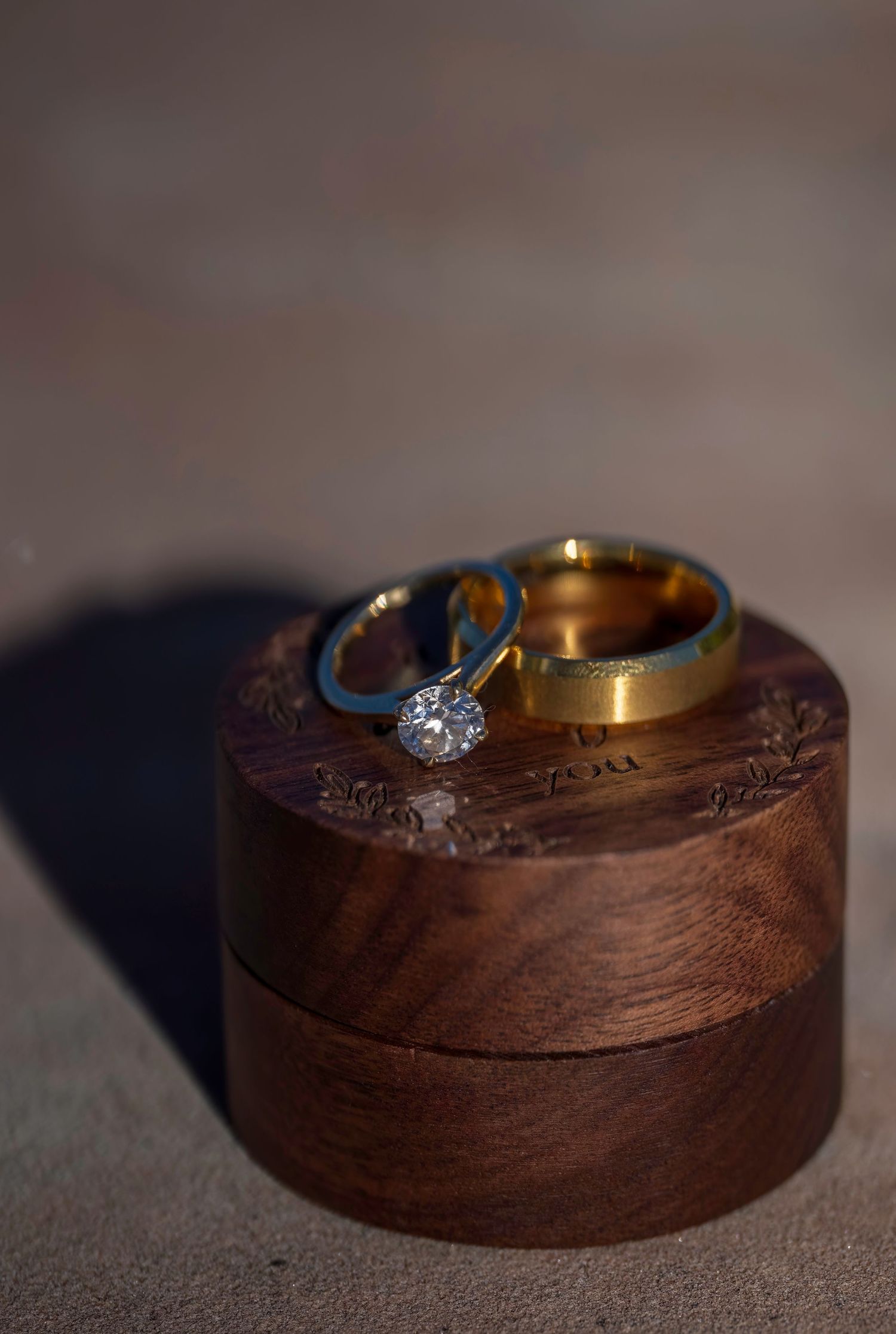 Two gold wedding bands and a diamond engagement ring displayed on a round wooden ring box.