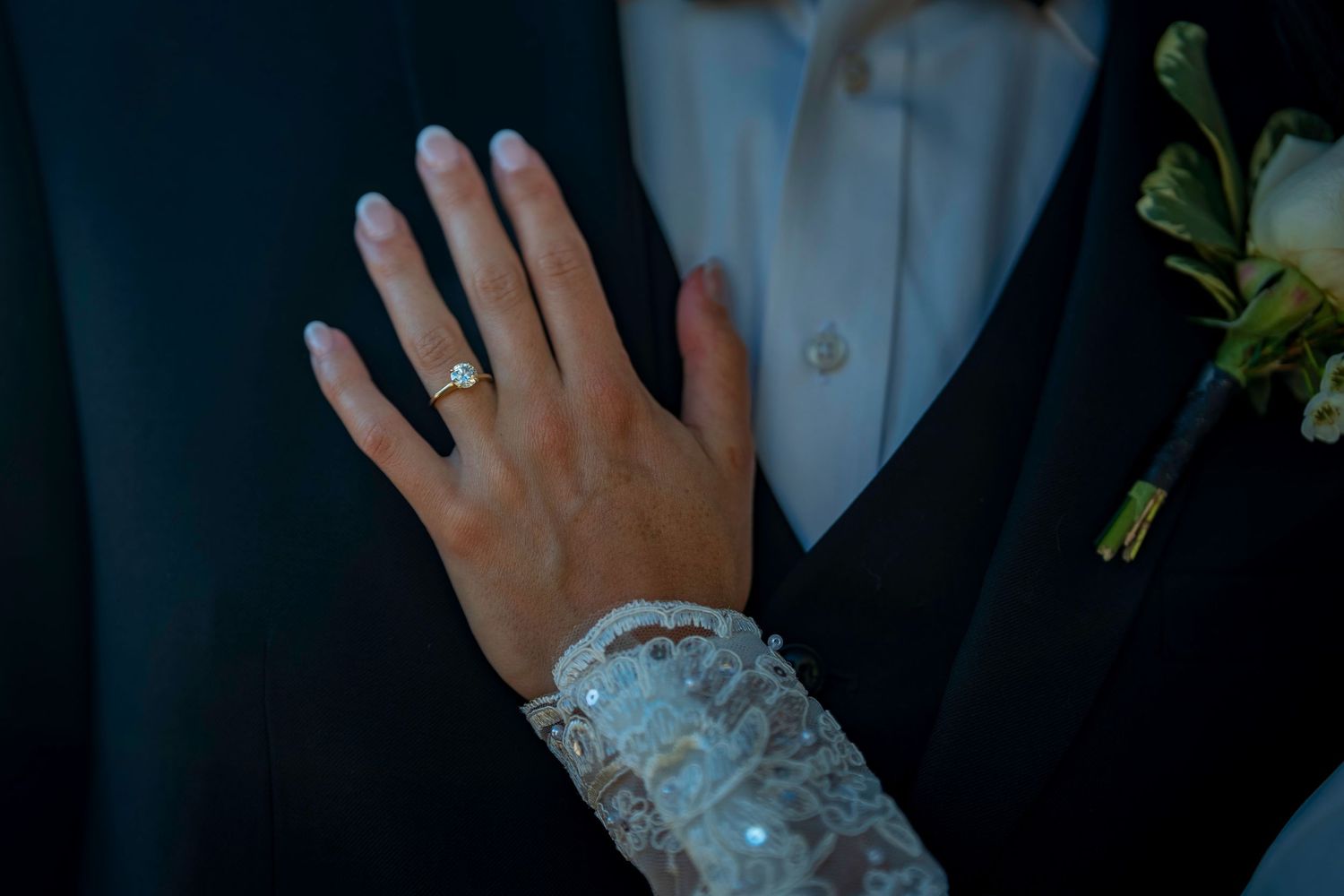 Close-up of bride's hand showing diamond engagement ring with lace sleeve detail.