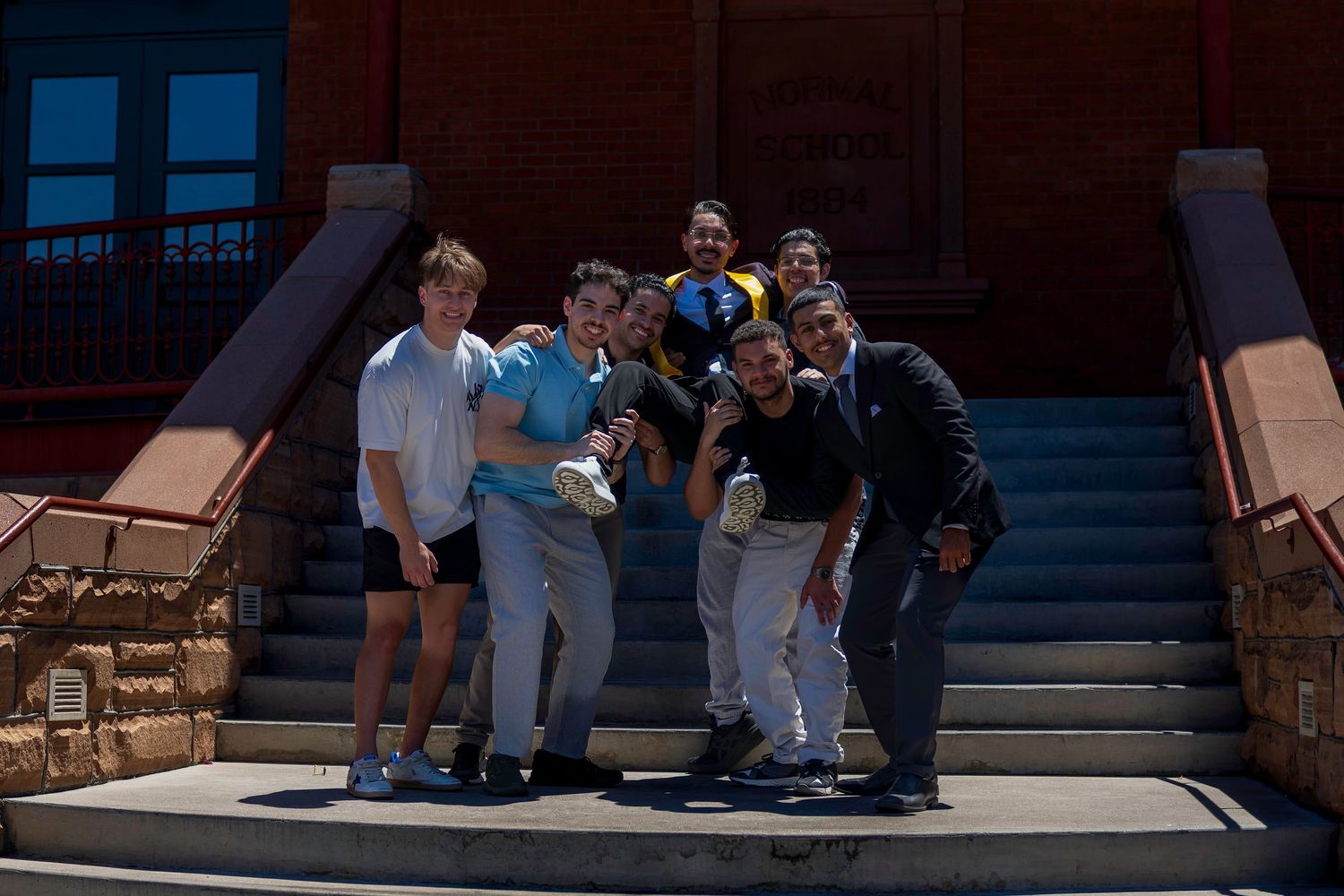 Group of young men posing together on steps outside a red brick building on a sunny day.