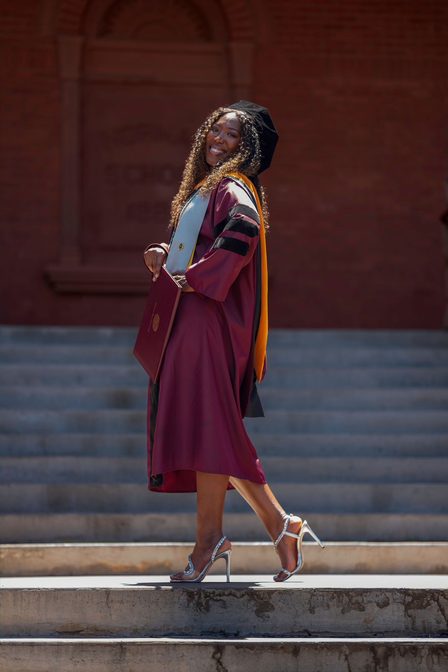 Smiling Black woman in maroon graduation gown and cap holding diploma on sunlit steps of brick building.