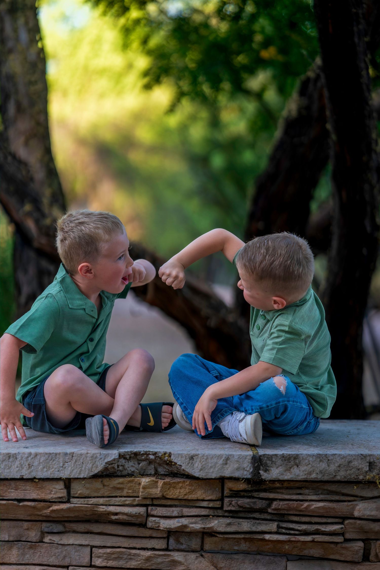 Two young twin boys in matching green shirts sit on a stone wall, playfully fist-bumping outdoors.