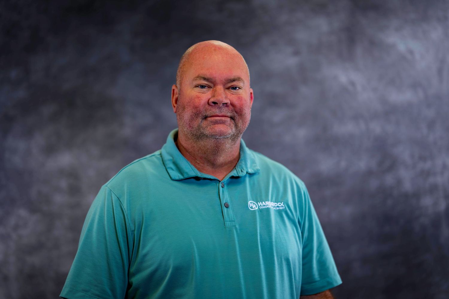 Professional headshot of employee in teal polo shirt against textured gray backdrop.