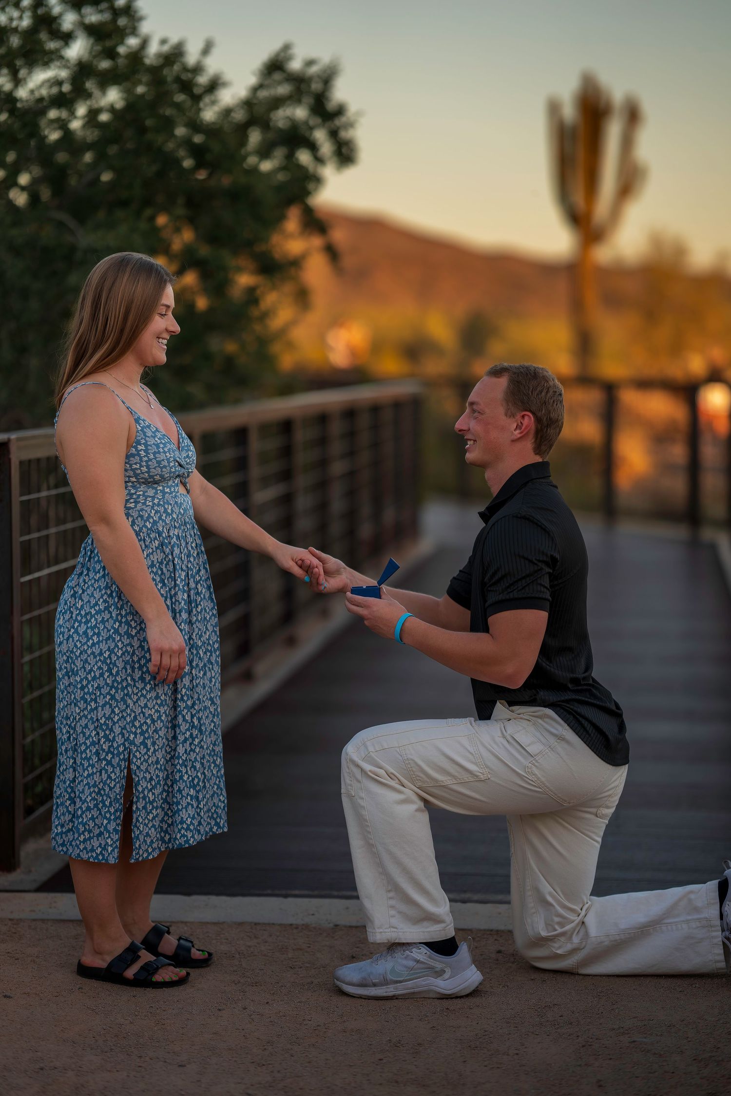 Man proposes on one knee to woman on a wooden bridge at sunset in Arizona desert landscape.