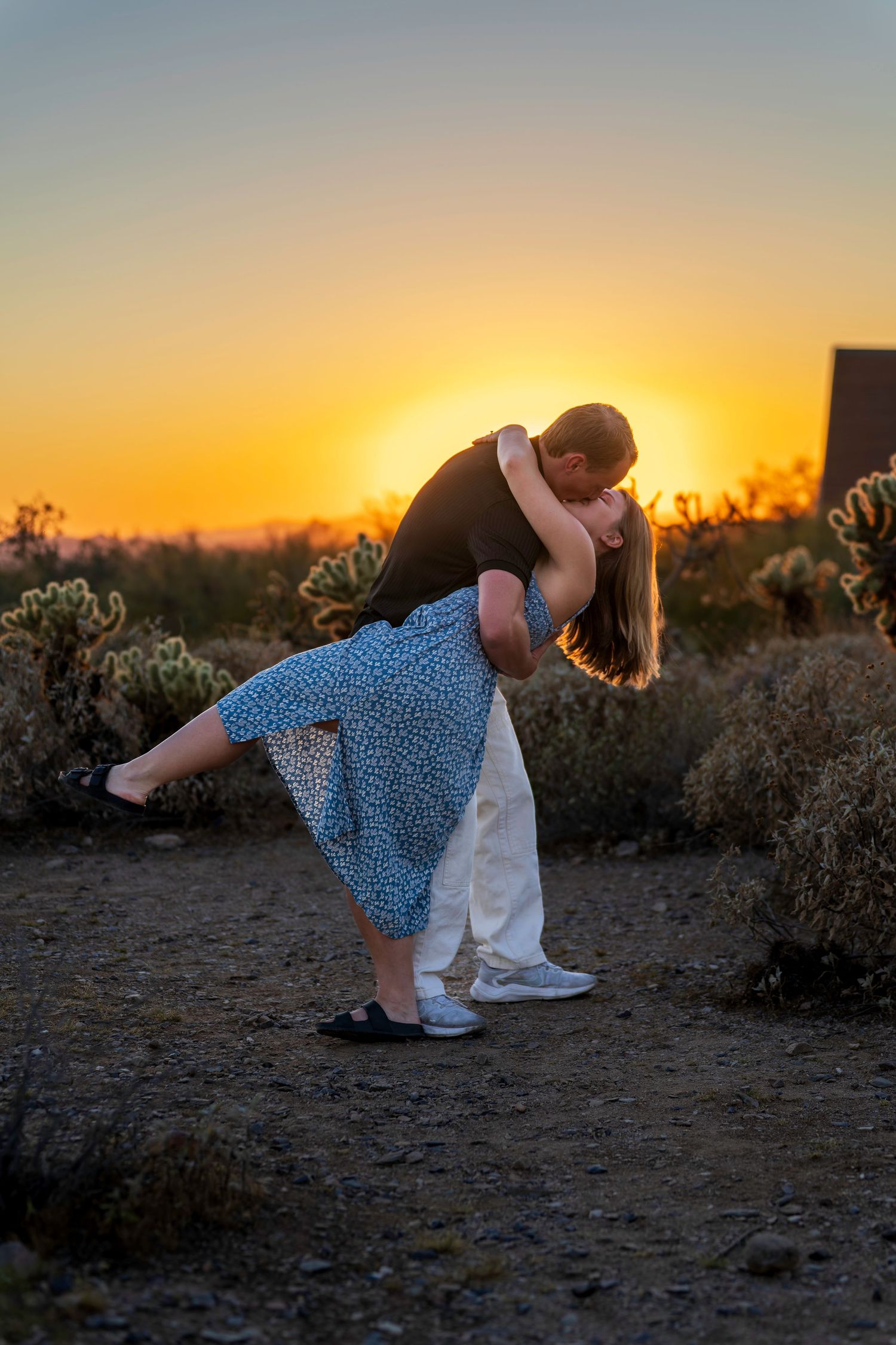 Couple sharing romantic dip kiss silhouetted against vibrant orange desert sunset with cacti in background.