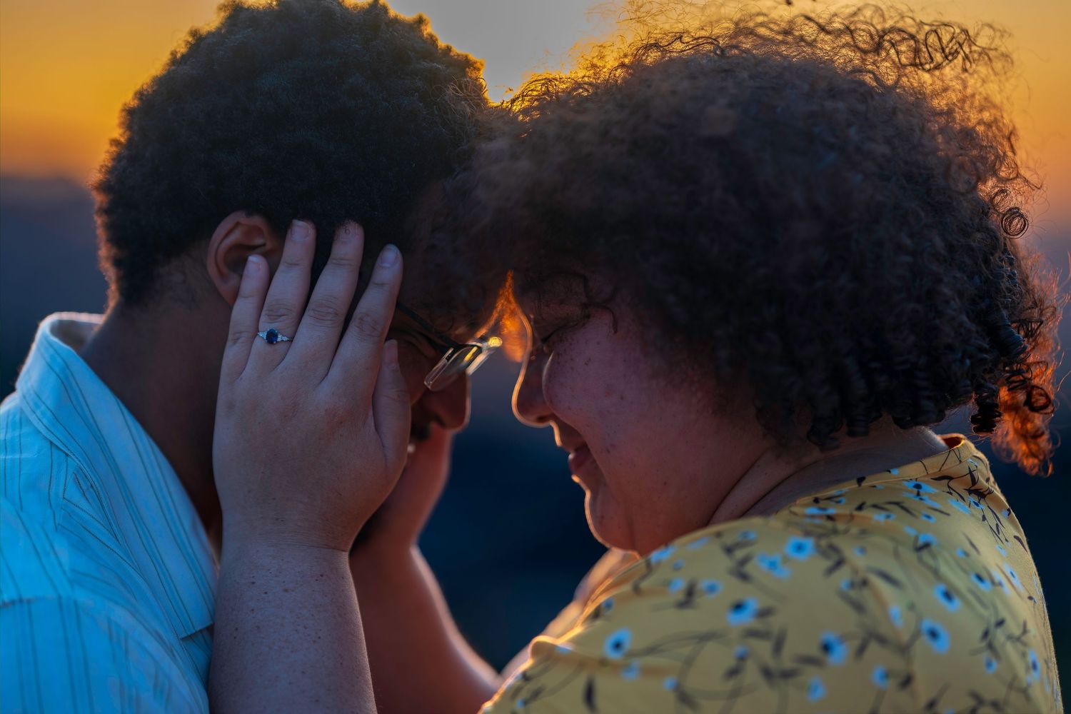 Two people with curly hair touch foreheads tenderly at sunset, sharing an intimate romantic moment outdoors.