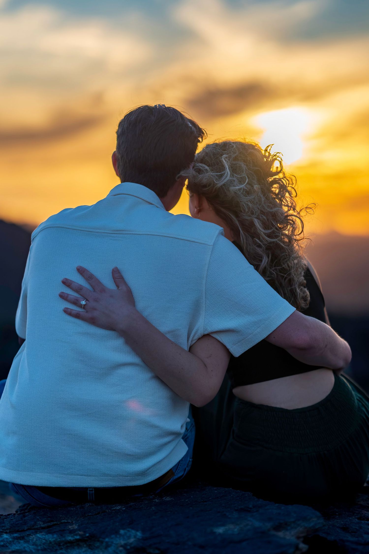 Couple sitting together arm in arm watching a vibrant golden sunset from a rocky overlook.