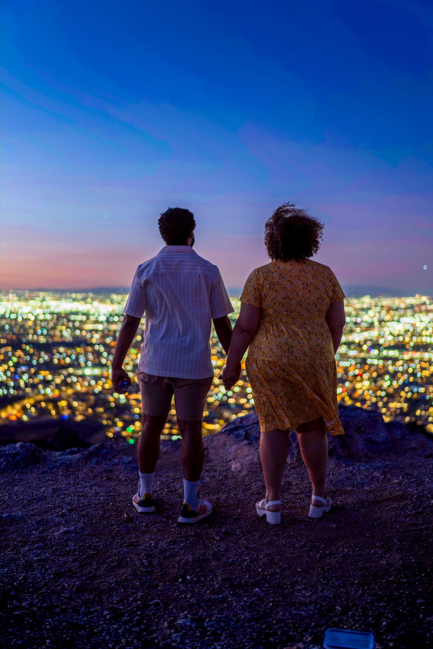 Couple holding hands overlooking breathtaking city lights from mountain overlook at night.