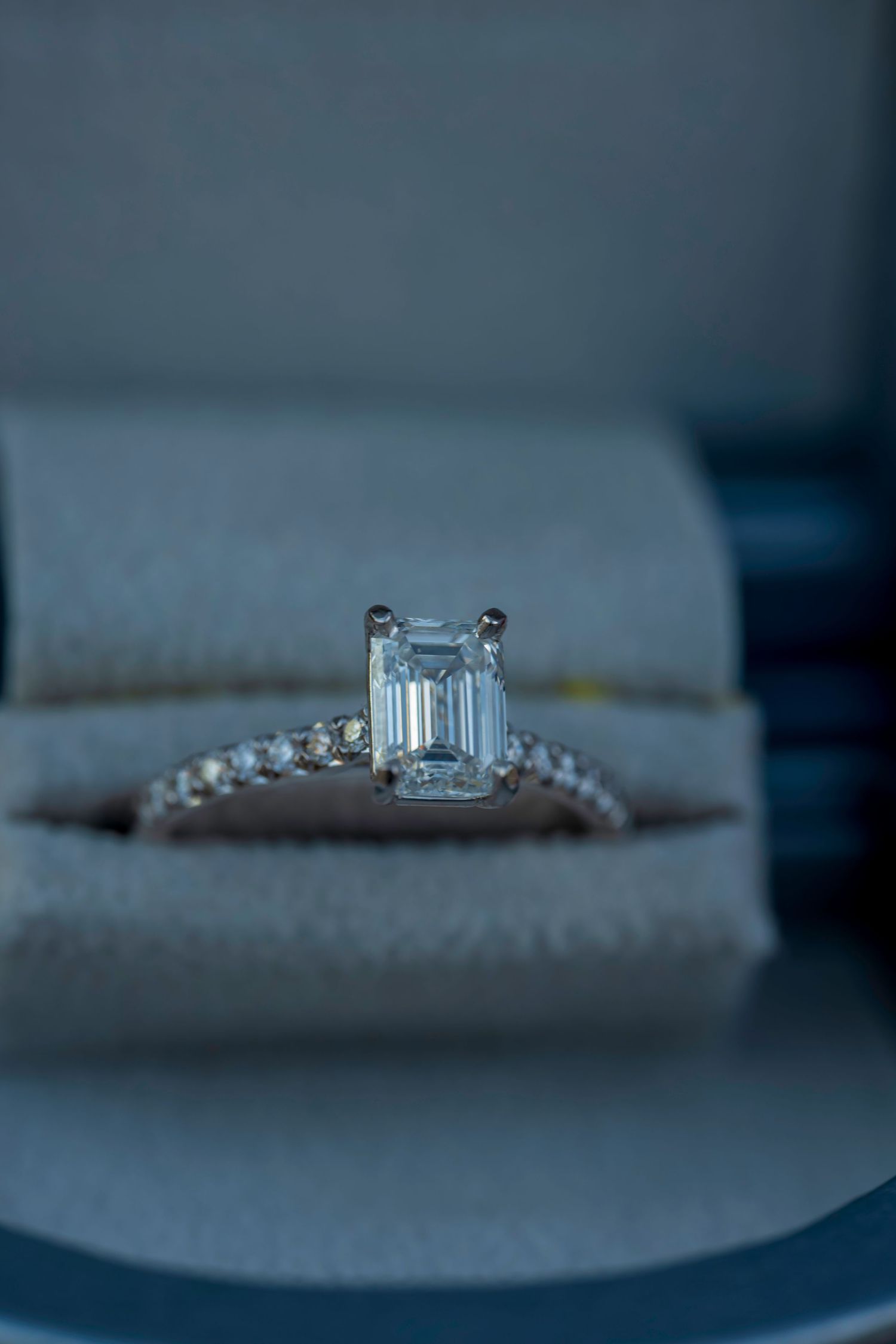Close-up of an emerald cut diamond engagement ring with pavé band displayed in a gray velvet ring box.