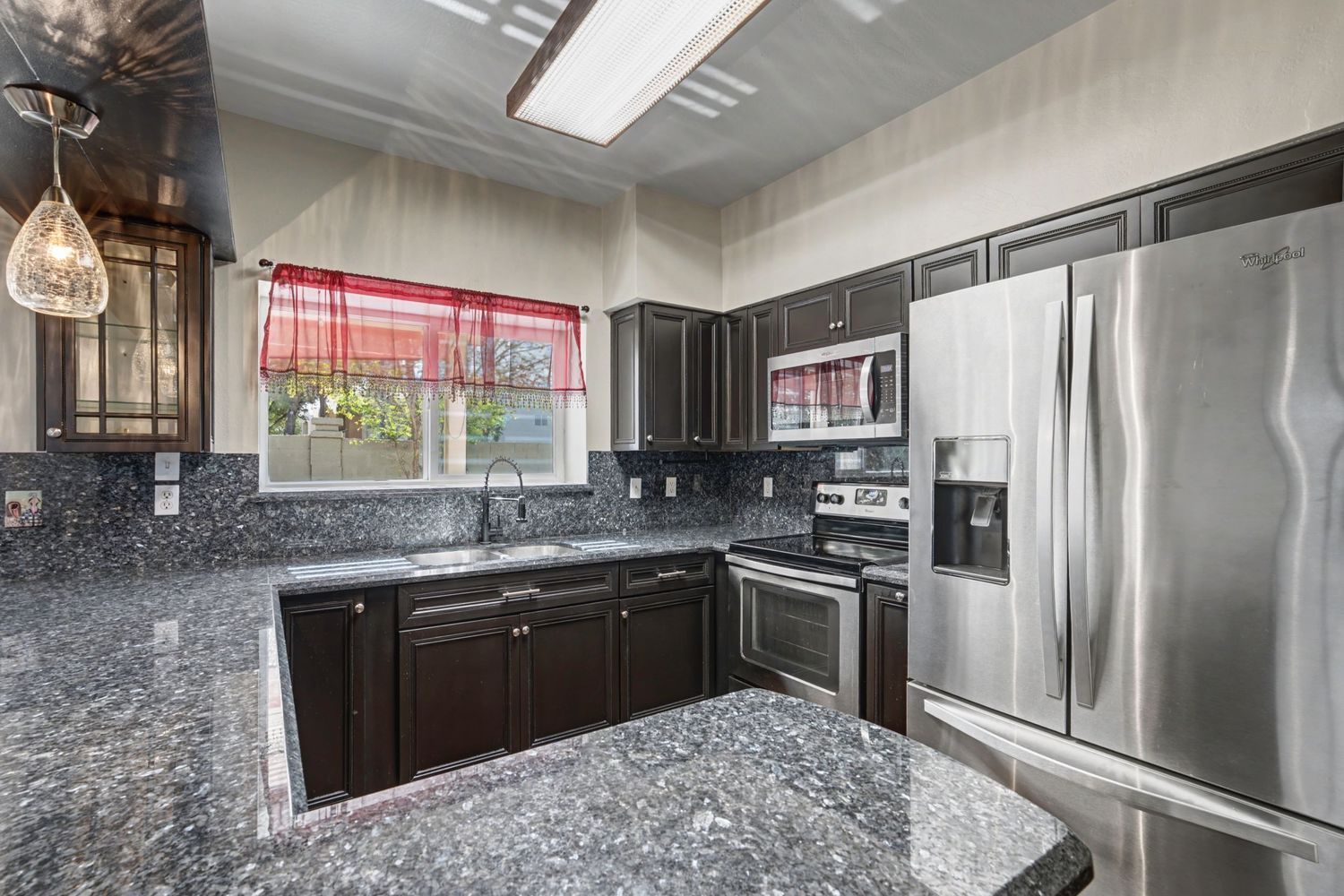 Dark espresso kitchen cabinets with granite countertops, stainless steel appliances, and red window valance.