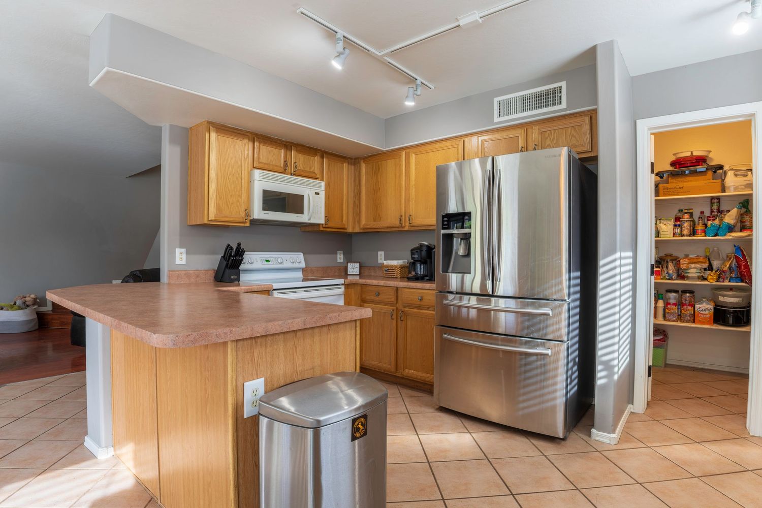 Modern kitchen with stainless steel refrigerator, oak cabinets, white appliances, tile floors, and a pantry visible in the background.