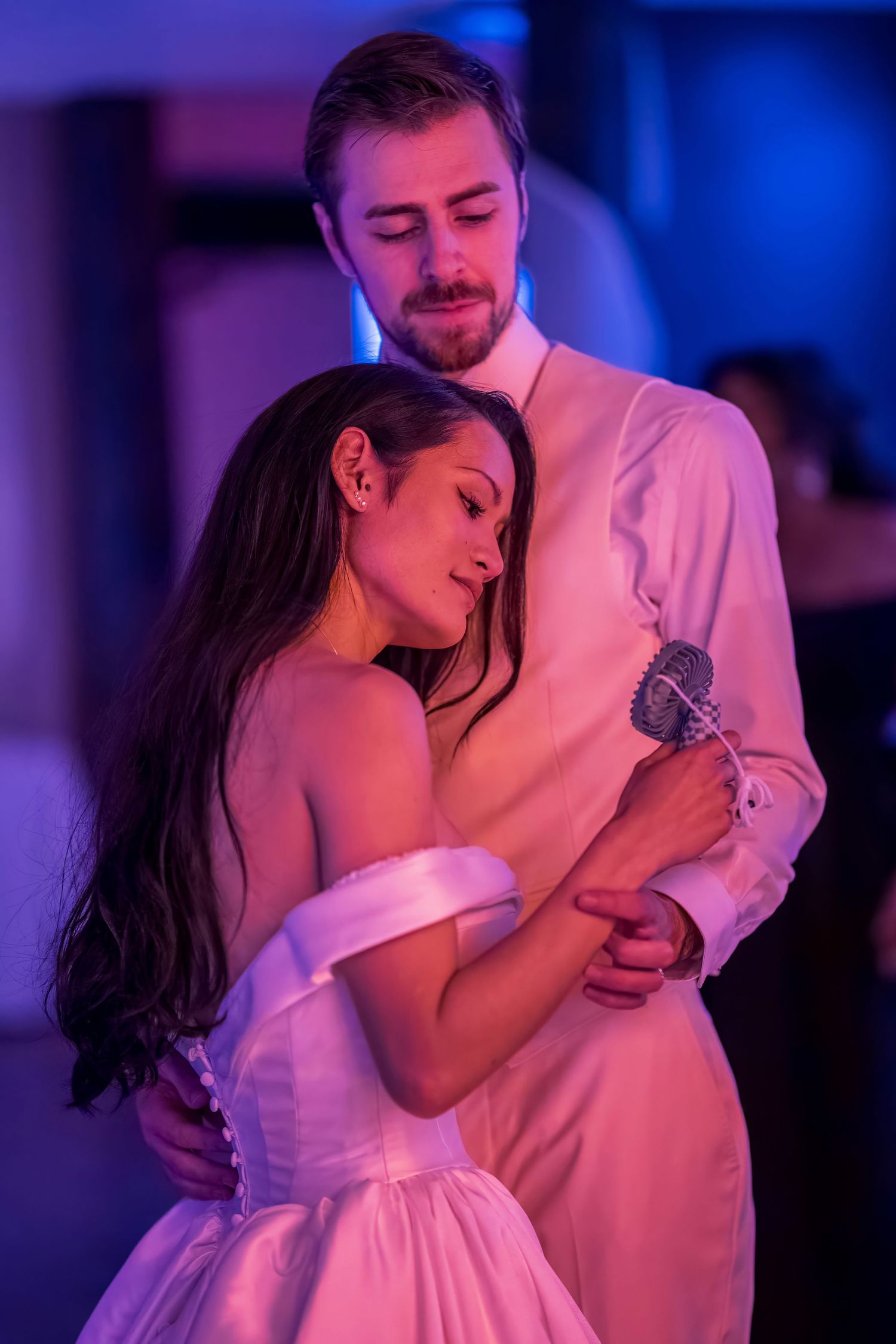 A couple embraces during a slow dance at their wedding reception with purple lighting.