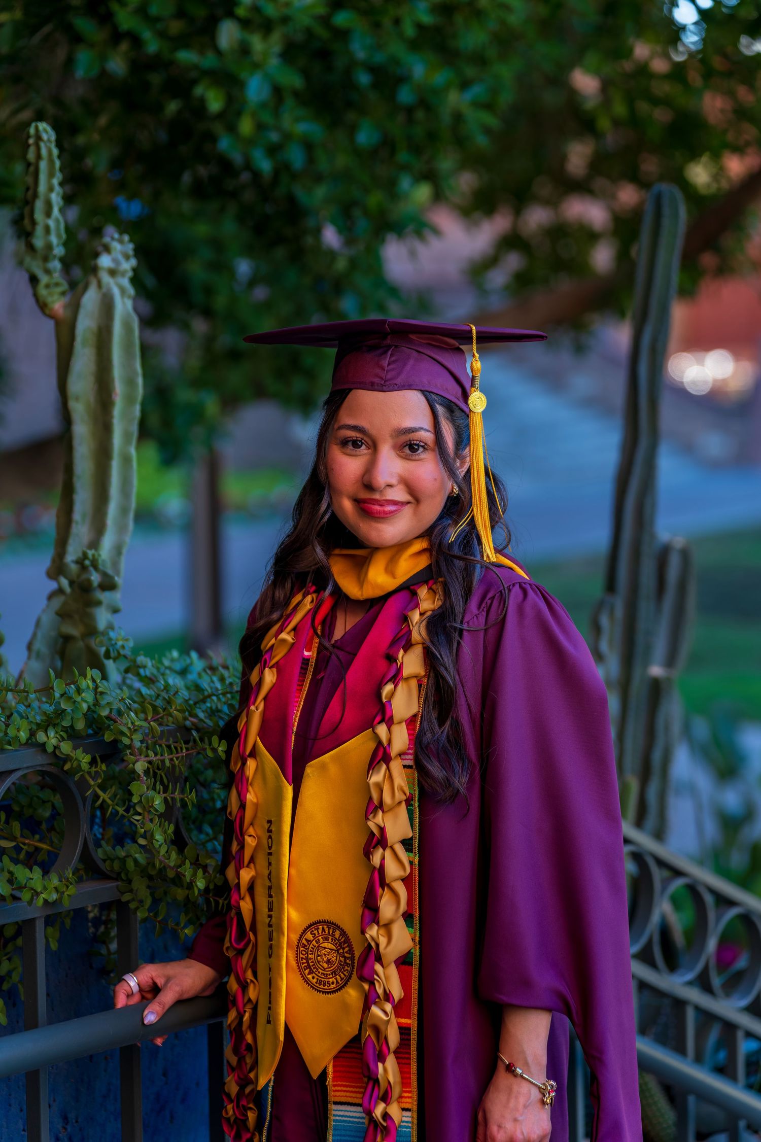 Graduate in maroon cap and gown stands before blue door at dusk.