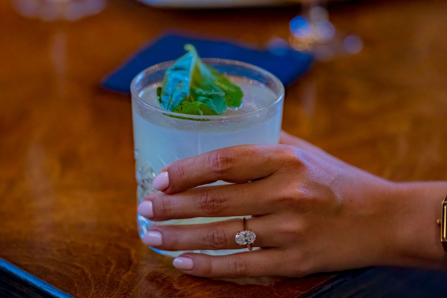 A hand wearing an engagement ring holds a blue cocktail garnished with mint.