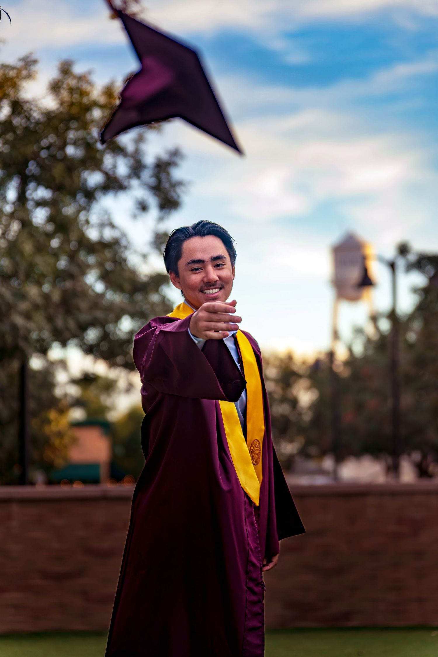 Graduate in maroon and gold cap and gown poses outdoors during sunset photo session.