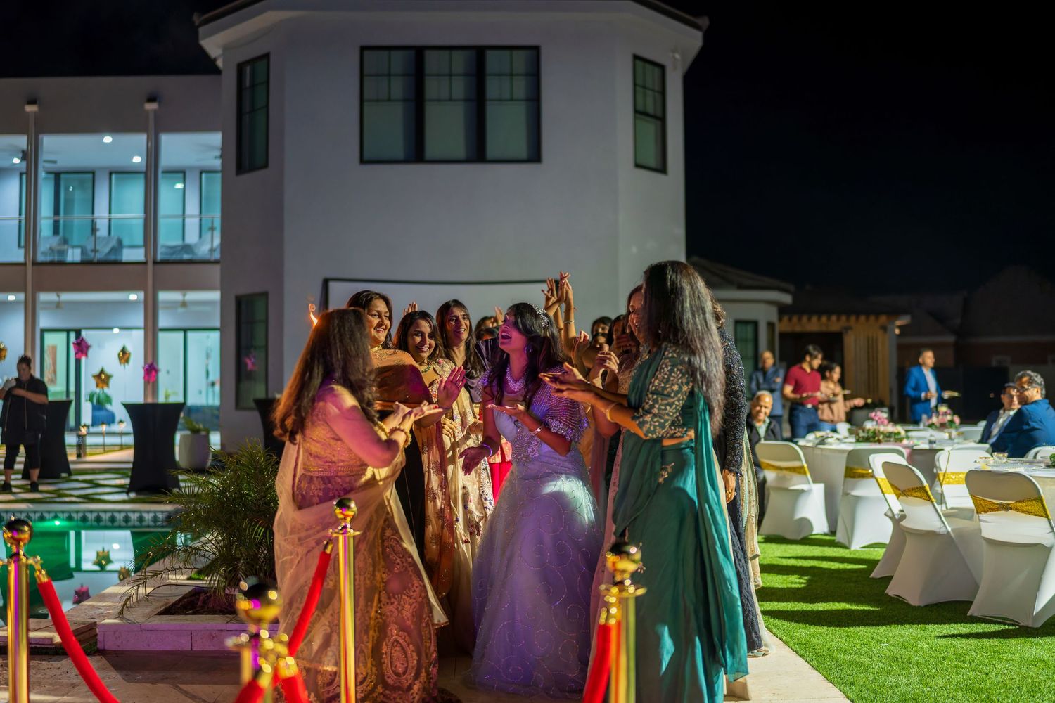 An outdoor evening celebration with guests in formal attire gathered under building lights.