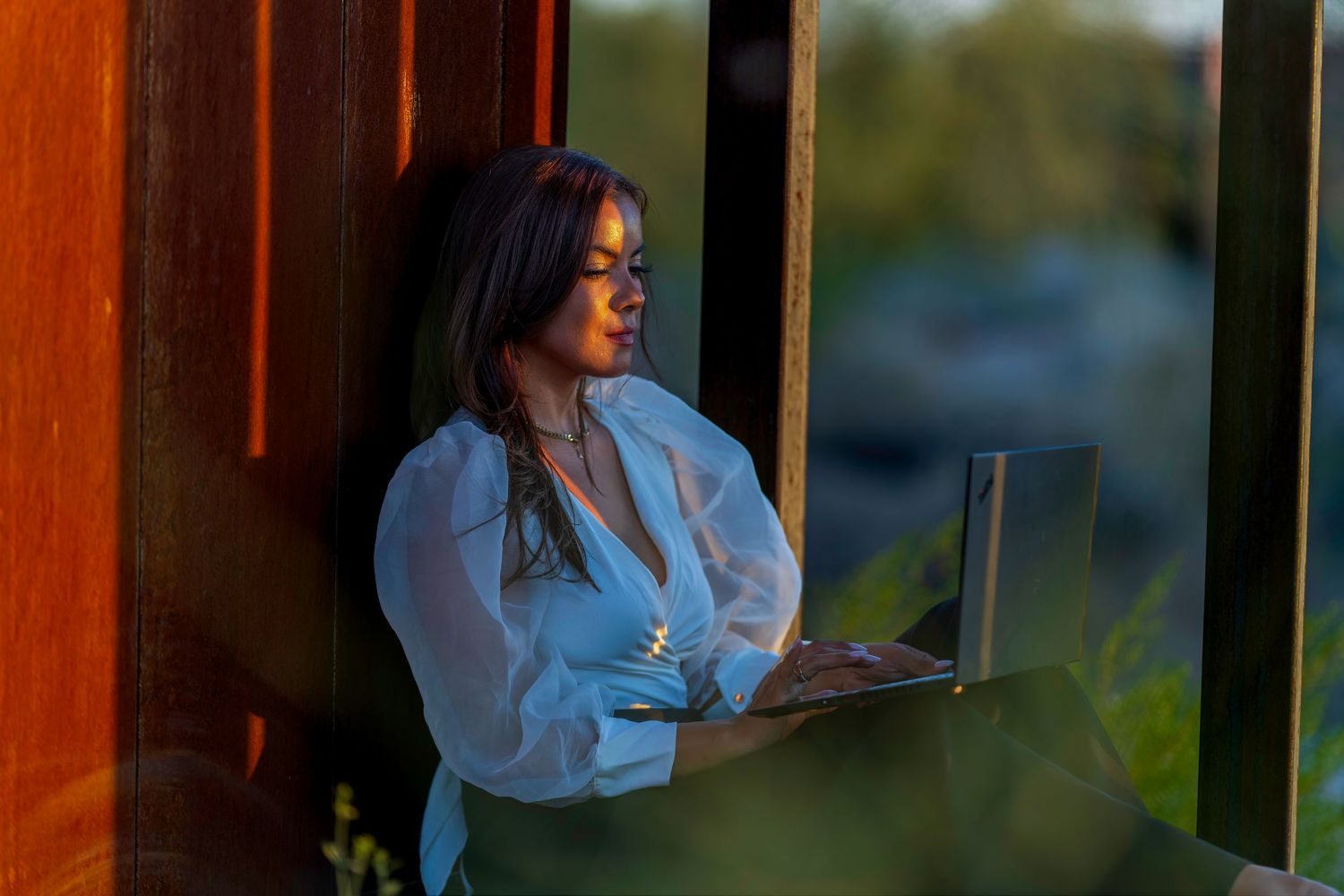 Woman in white blouse working on laptop outdoors in warm golden hour light, leaning against wooden wall.