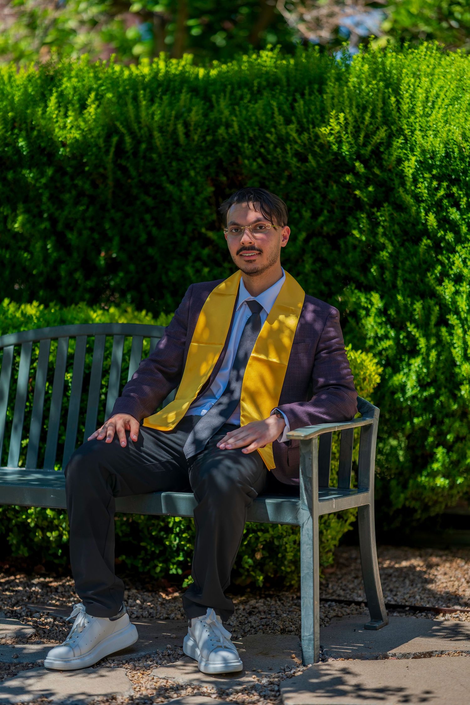 Graduate in maroon suit with gold stole sits on bench surrounded by lush greenery.