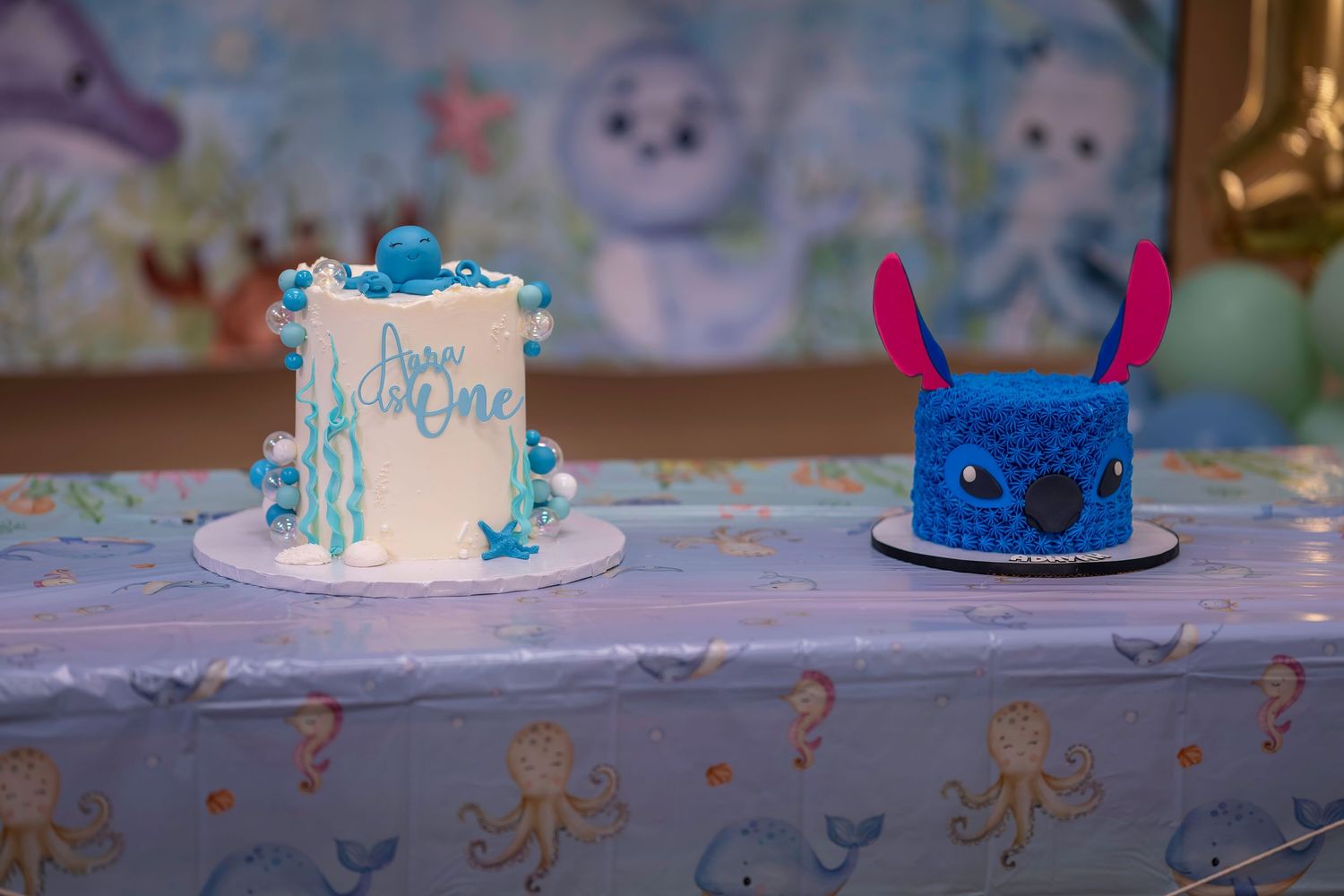 Two birthday cakes on a ocean-themed table, one white with blue writing and one blue Stitch-shaped smash cake.