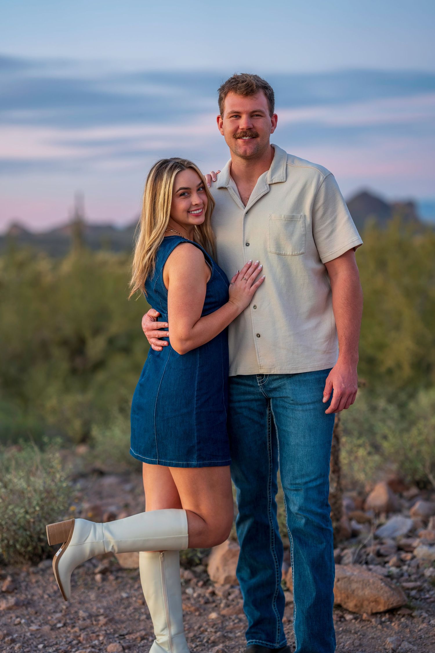Couple posing together at sunset in desert landscape, woman in denim dress and white boots, man in cream shirt and jeans.
