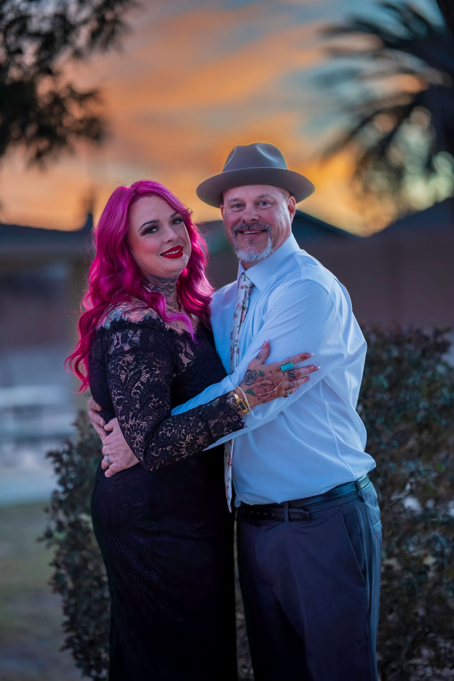 Couple embracing at sunset, woman with pink hair in black lace dress, man in white shirt and gray fedora.