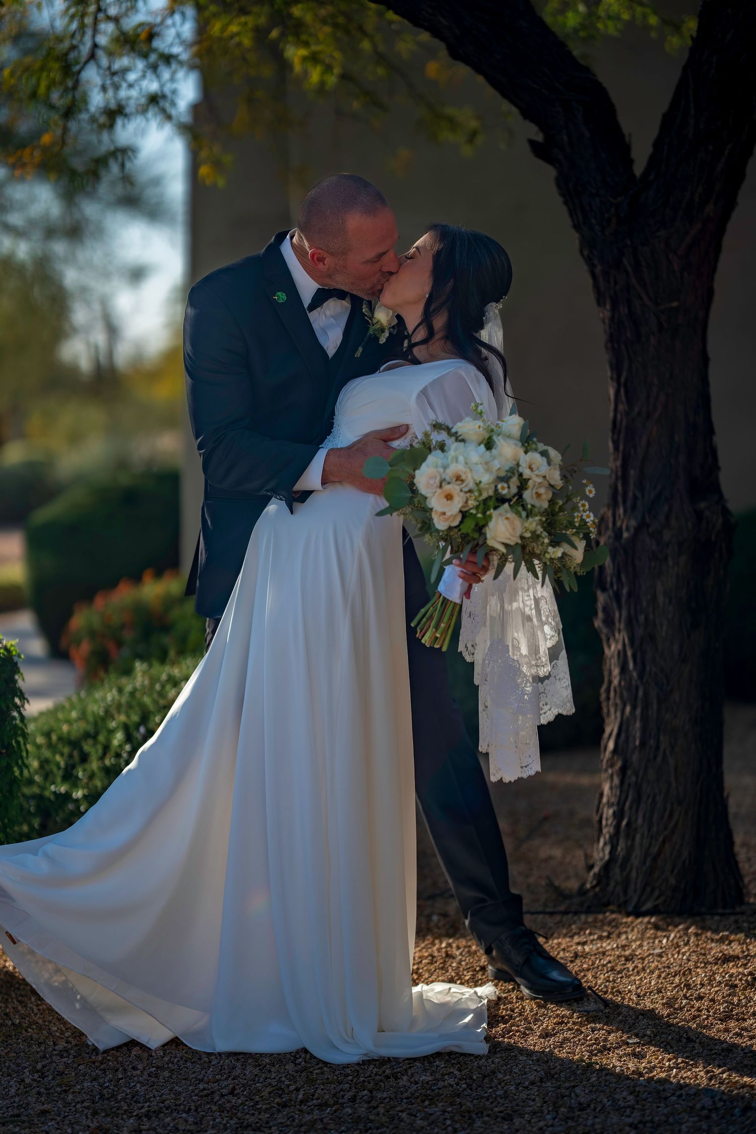 Bride and groom sharing a romantic kiss outdoors while bride holds white floral bouquet.
