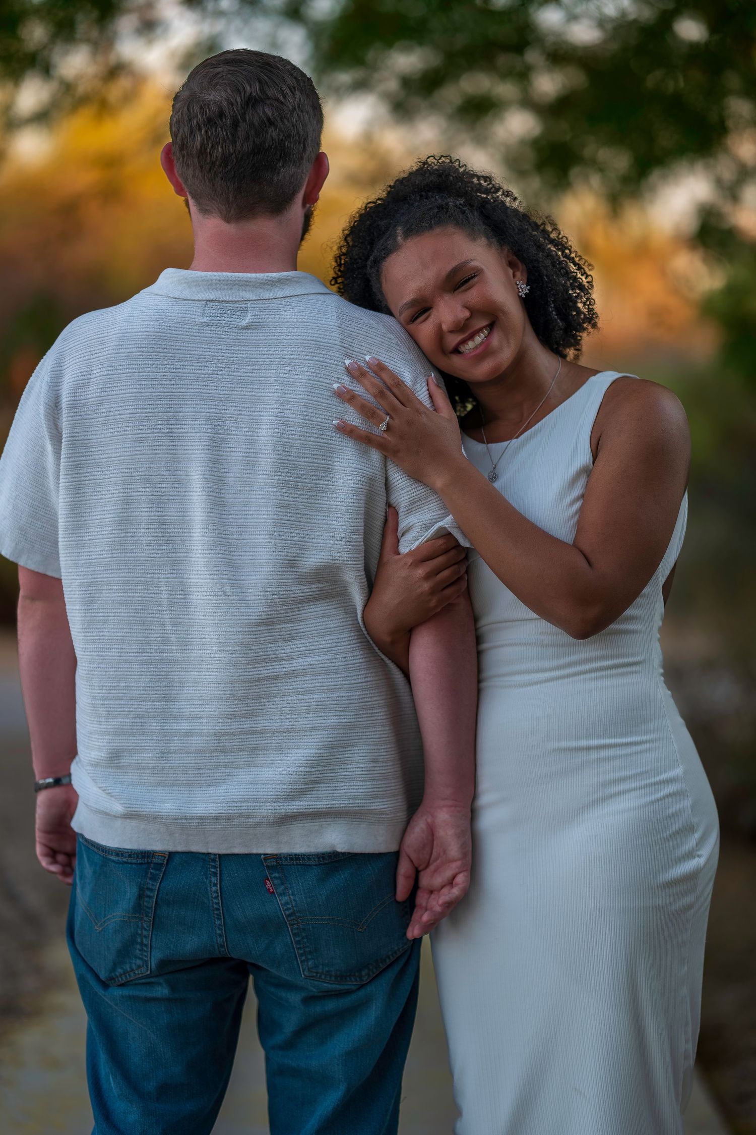 A smiling woman in a white dress embraces a man from behind during a golden hour outdoor engagement photo.