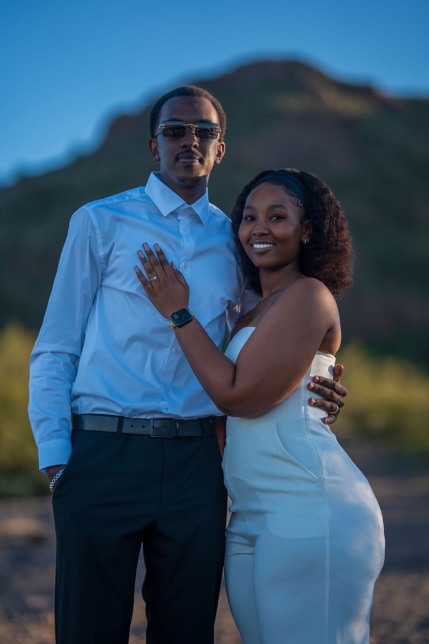 A couple poses outdoors in desert landscape, woman in white dress and man in white shirt with sunglasses.