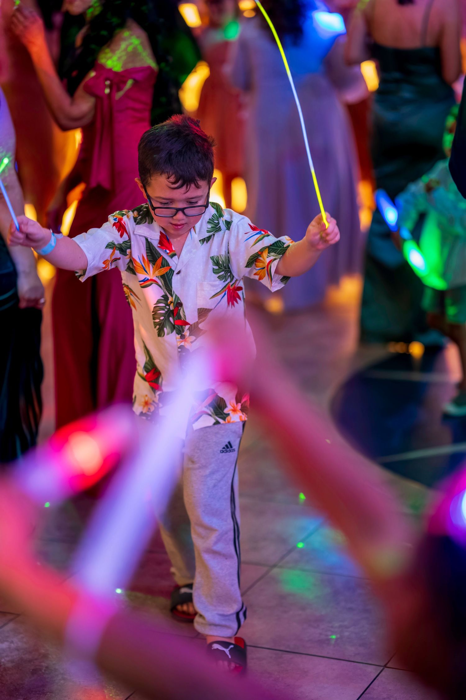 Young boy in floral shirt dancing with glow sticks at a colorful party event.