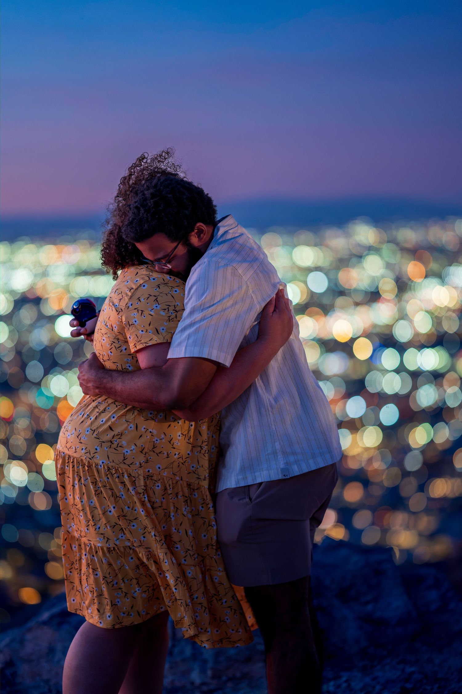 Couple sharing emotional hug at night with city lights bokeh glowing in background behind them.